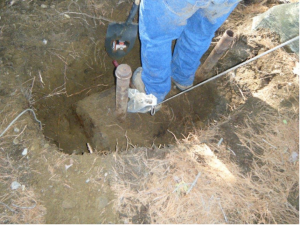 A person holding a shovel standing in a shallow hole dug around an underground oil tank filler pipe.