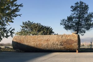 A removed, old rusty underground oil tank sits out in the open of a parking area with trees behind it.